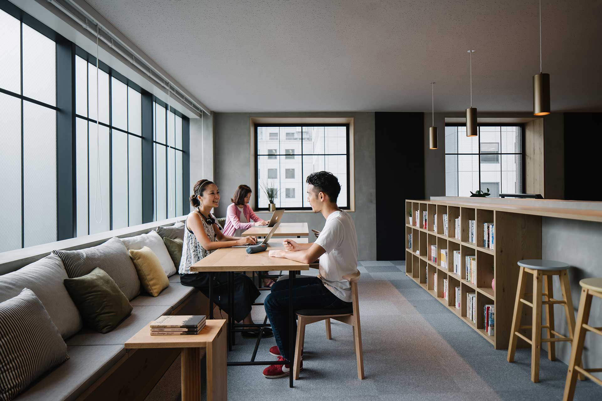 Three people sit in a minimal, clean workspace with cubby bookshelves and window benches.